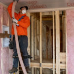 Worker on stilts using a hose to install blown-in insulation in a ceiling.