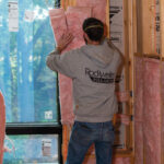 Rockweiler Insulation worker installing batt insulation in a wall next to a window.
