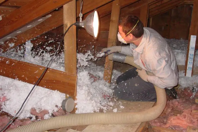 Worker blowing loose-fill insulation in an attic wearing a respirator mask.