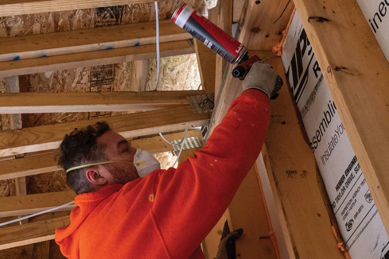 Worker in respirator applying can foam to seal gaps along roof rafters during new construction.