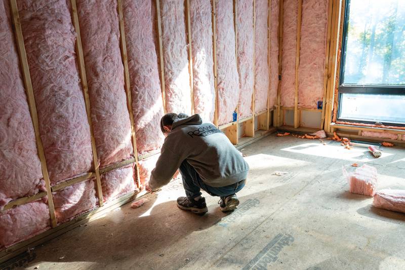 Insulation worker crouching to fit batt insulation along the base of a framed exterior wall.