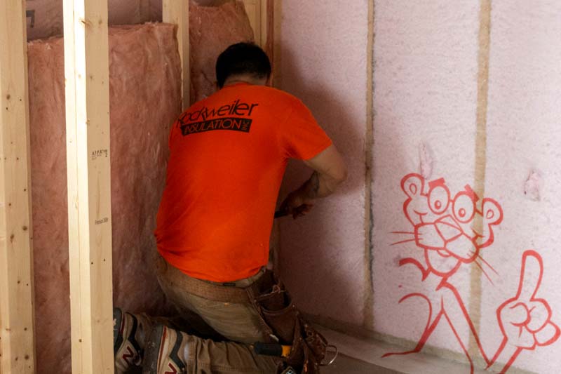 Rockweiler Insulation installer fitting pink fiberglass batt insulation into wood-framed wall cavities in Madison, WI.